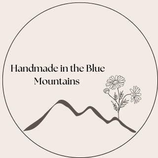 Minimalist circular design with a simple mountain line and flowers on the right, with the text 'Handmade in the Blue Mountains' centered above.