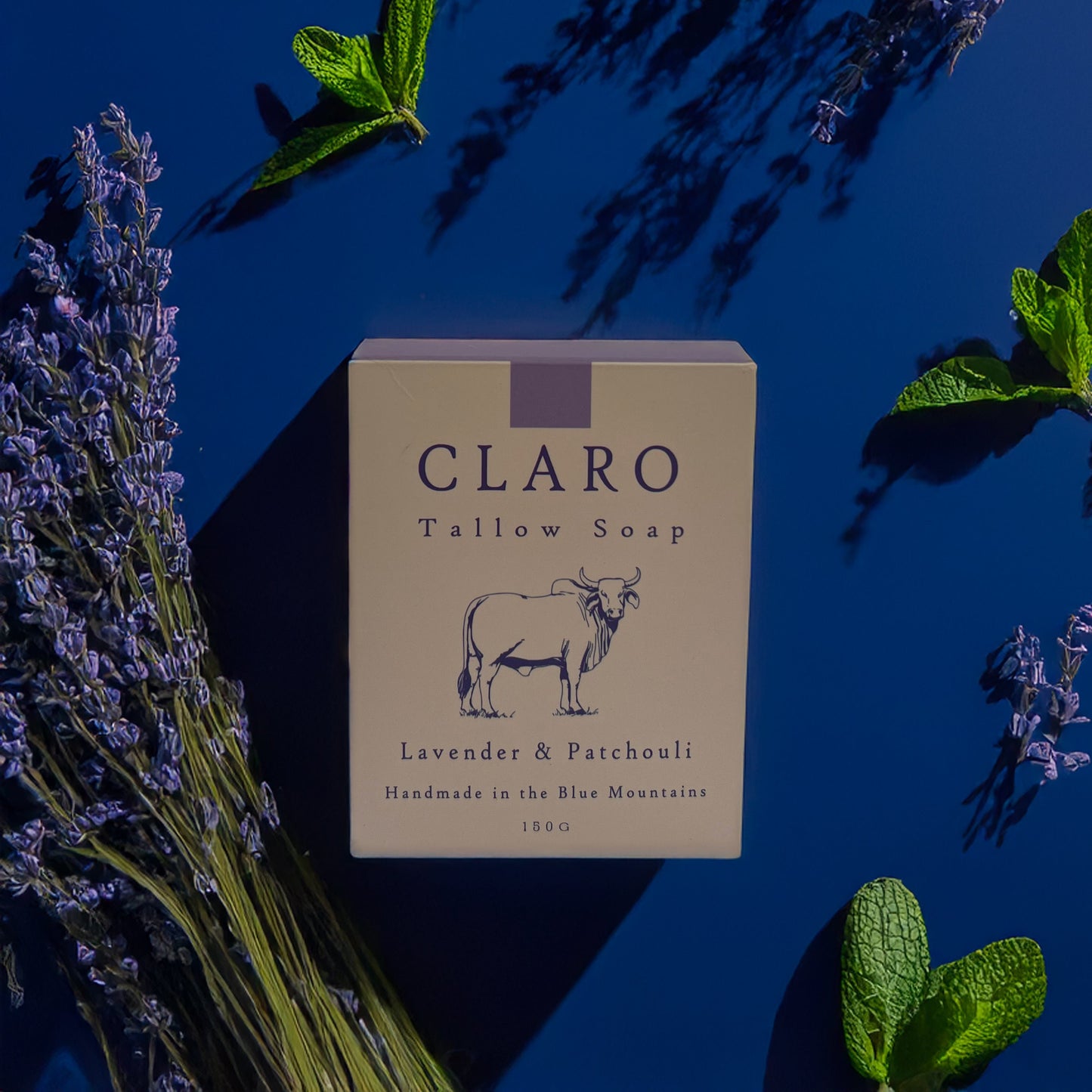 Claro Tallow Soap box labeled 'Lavender & Patchouli,' surrounded by sprigs of lavender and mint leaves on a dark blue background.