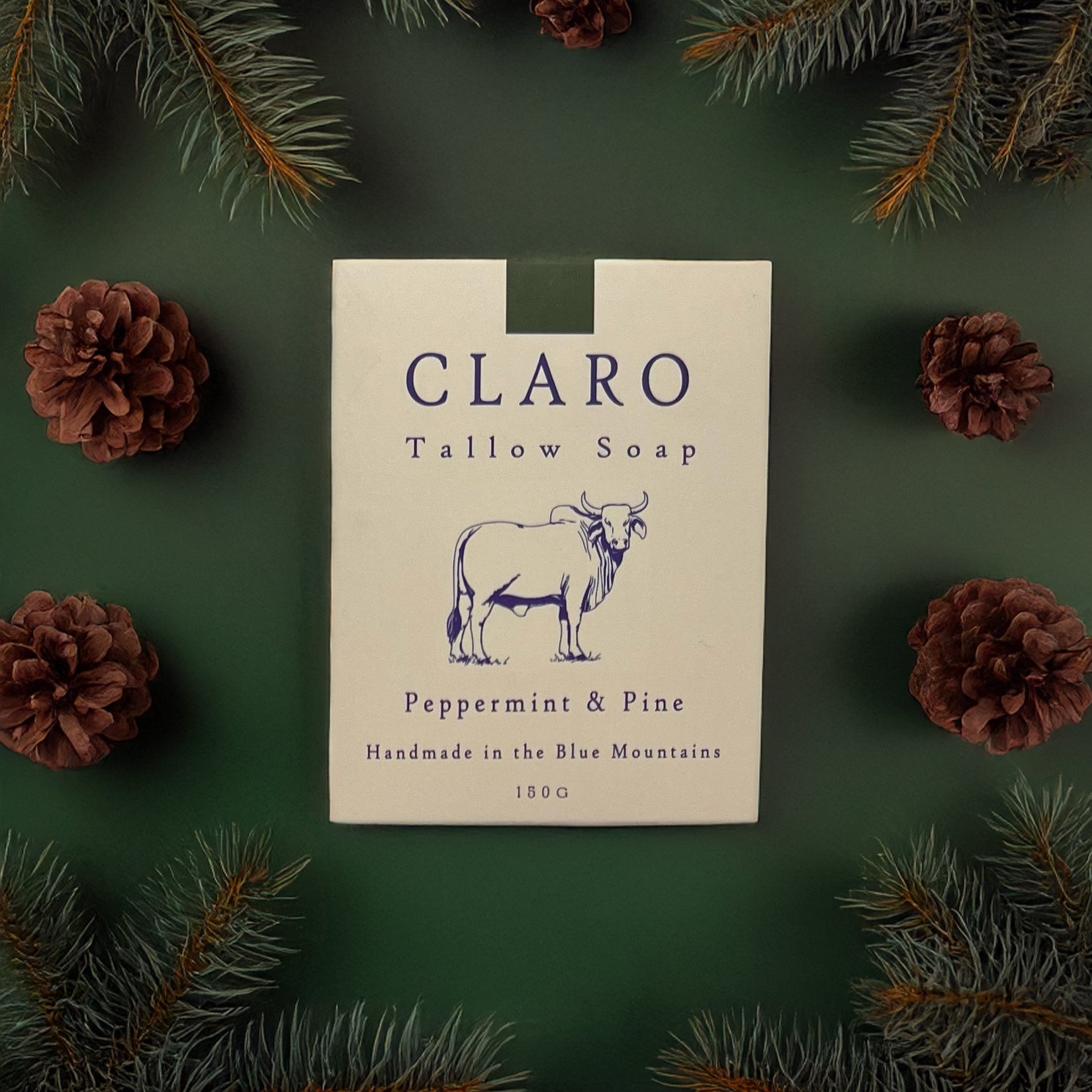 Claro Tallow Soap box labeled 'Peppermint & Pine,' surrounded by pine branches and pinecones on a green background.