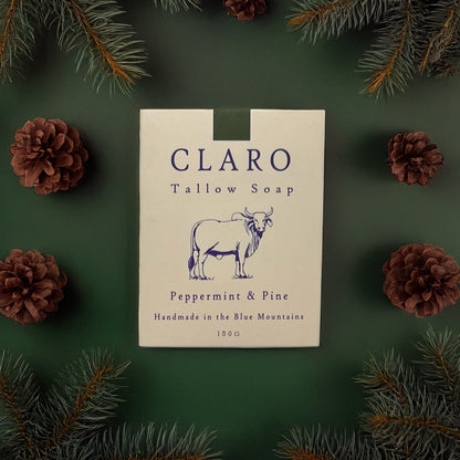 Claro Tallow Soap box labeled 'Peppermint & Pine,' surrounded by pine branches and pinecones on a green background.